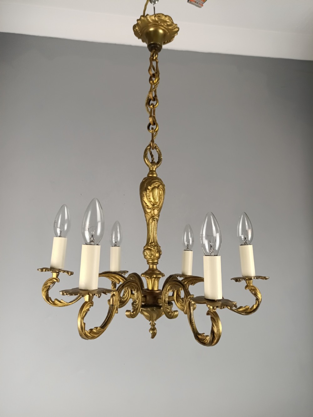 A French gilt brass six-arm chandelier featuring scrolled acanthus leaf branches, cream candle sleeves, and an ornate central column. View VI