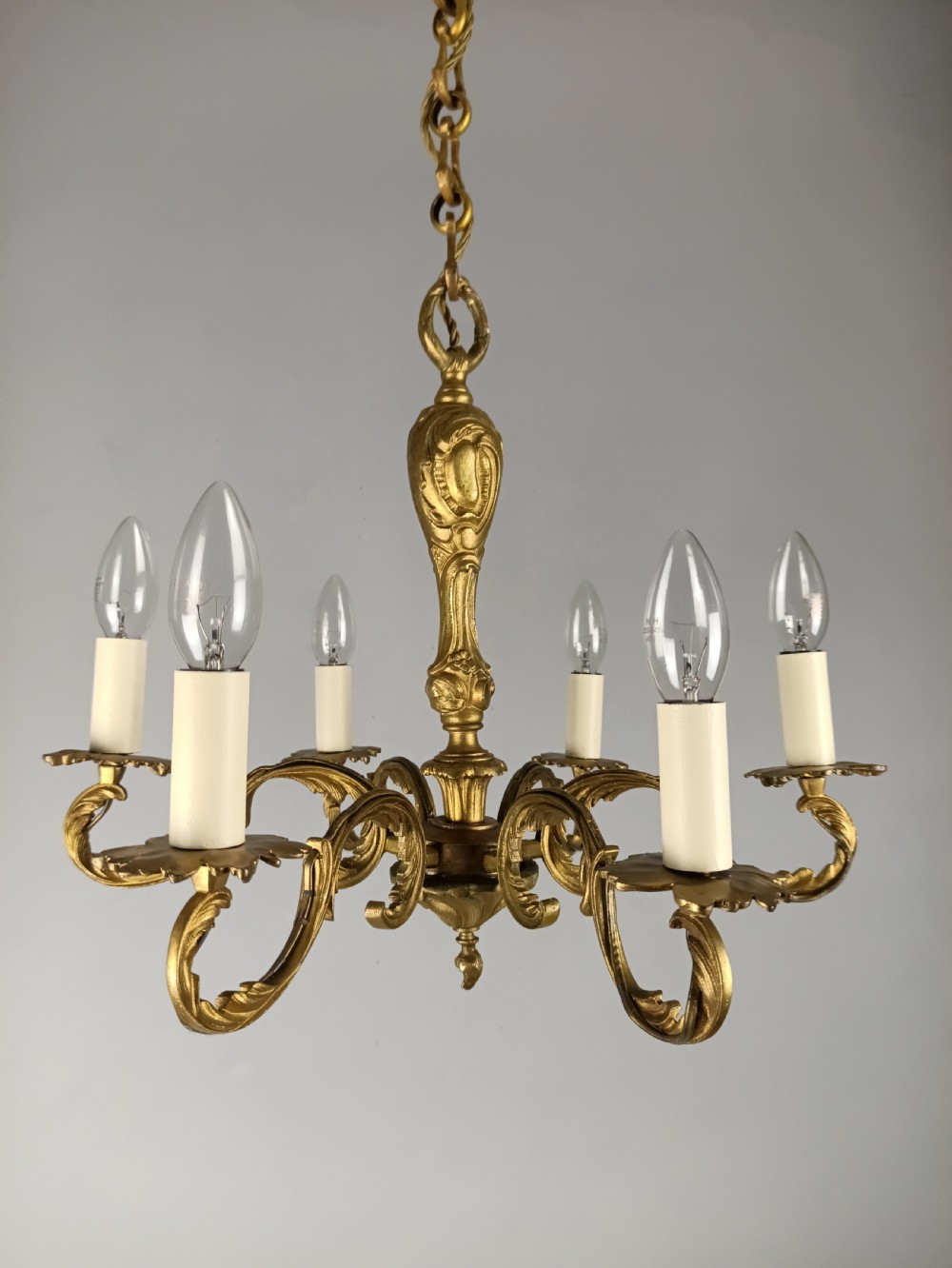 A French gilt brass six-arm chandelier featuring scrolled acanthus leaf branches, cream candle sleeves, and an ornate central column. View X