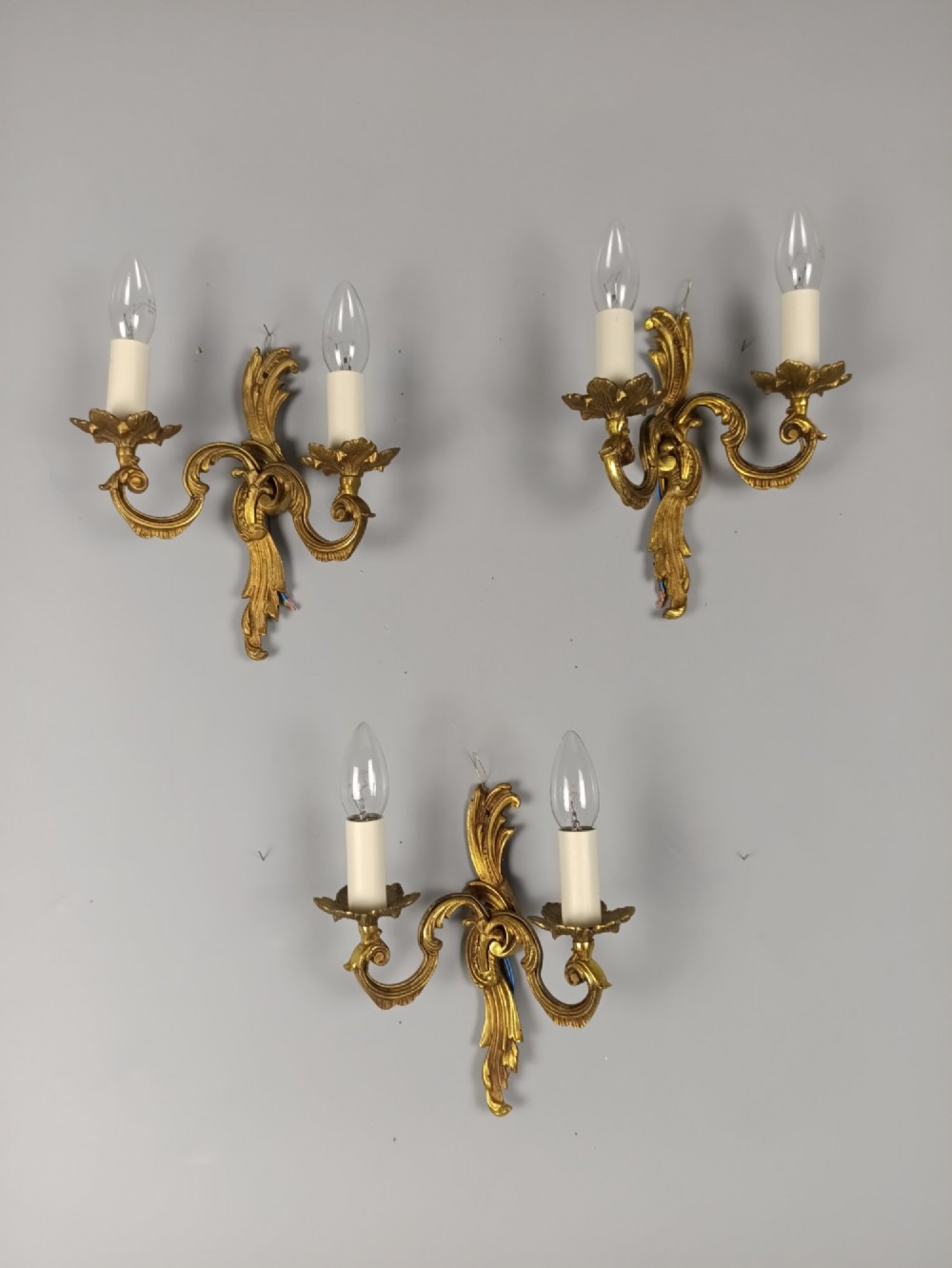 Set of three French gilt metal twin-arm wall sconces featuring acanthus leaf motifs and cream candle-style bulb sleeves.