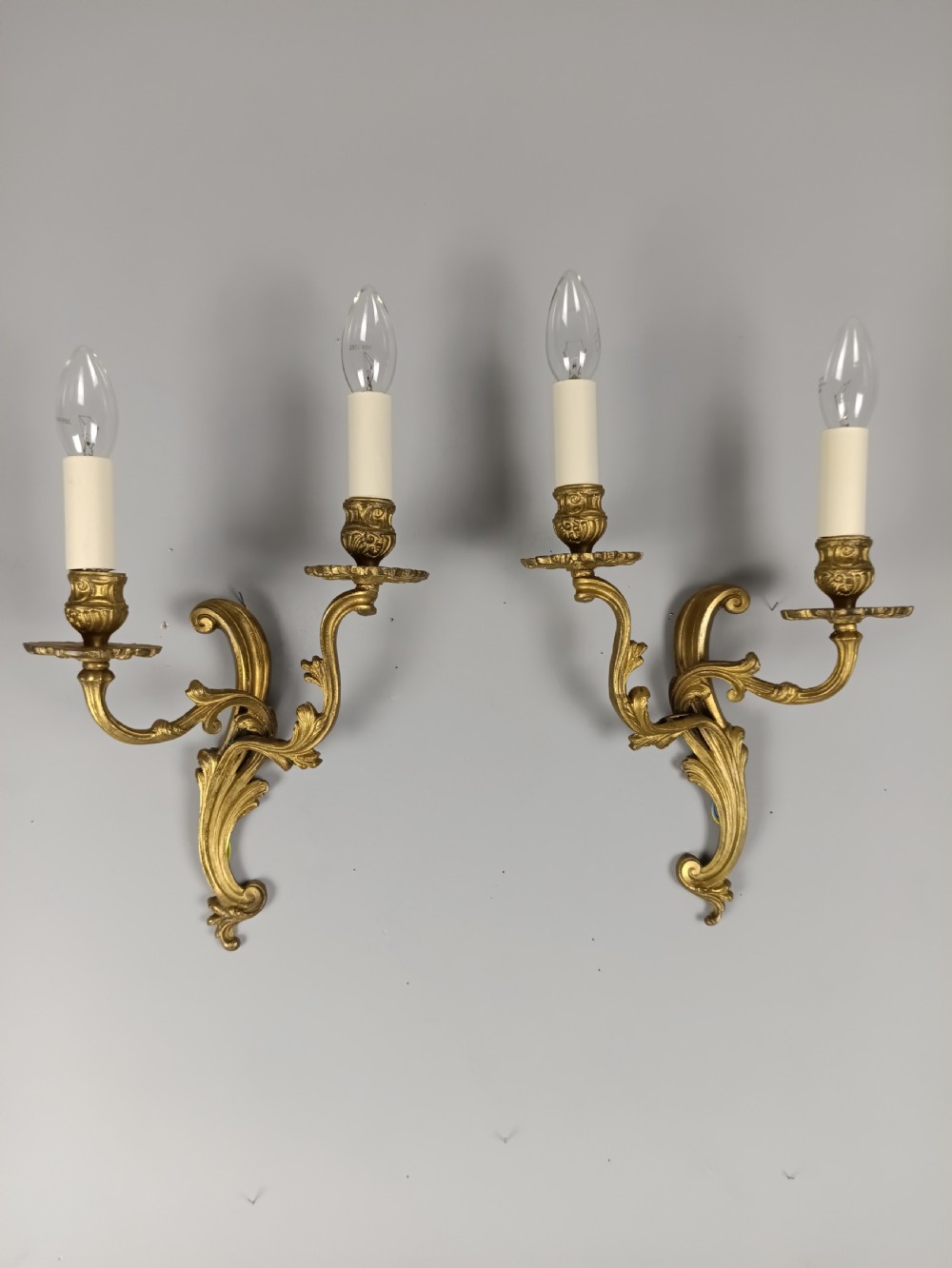 A pair of French gilt brass two-arm wall sconces featuring ornate acanthus leaf scrollwork and faux candle sleeve fittings.