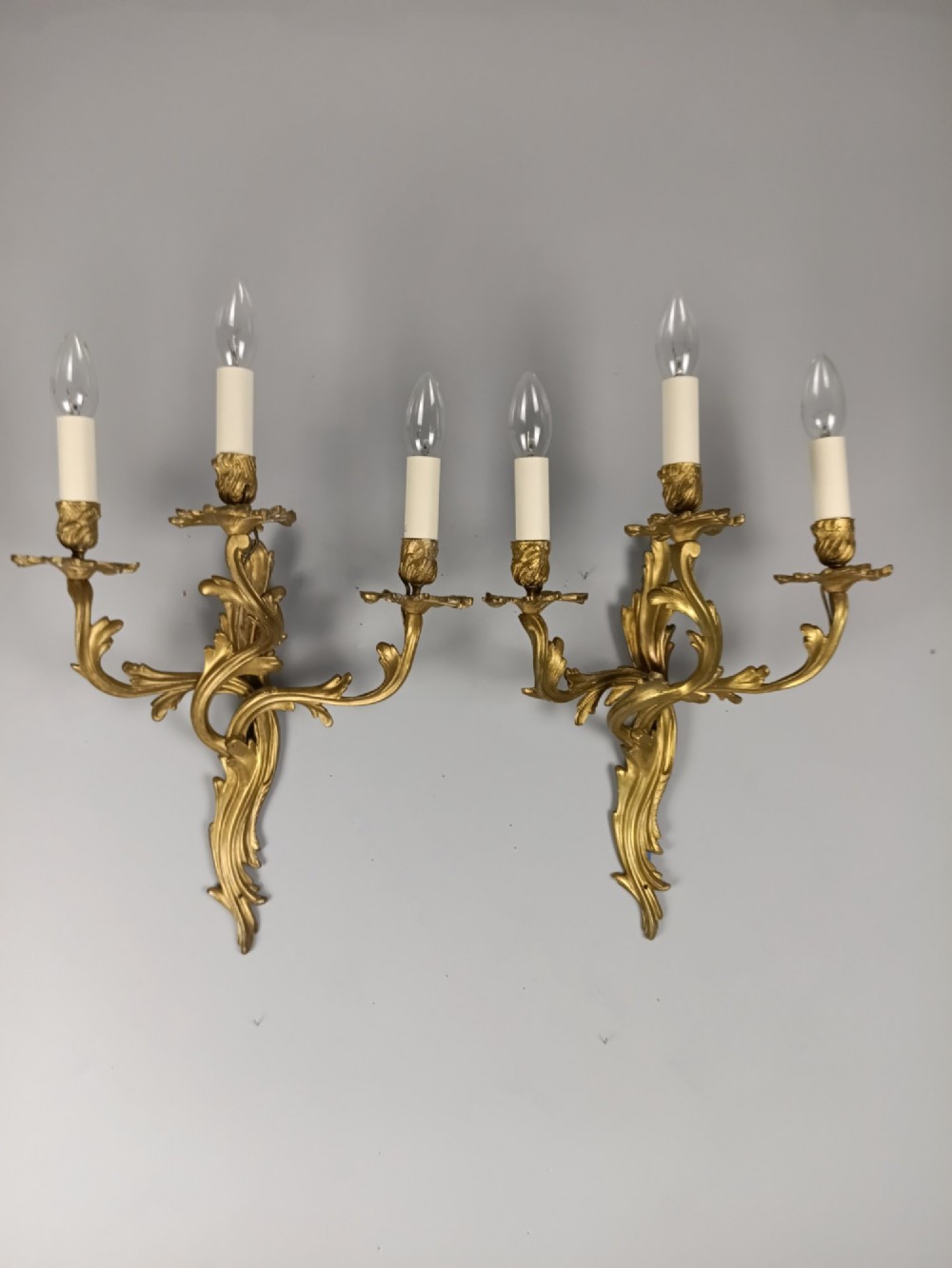 A pair of antique French Rococo gilded bronze triple-arm wall sconces featuring acanthus leaf scrollwork and candle sleeves.