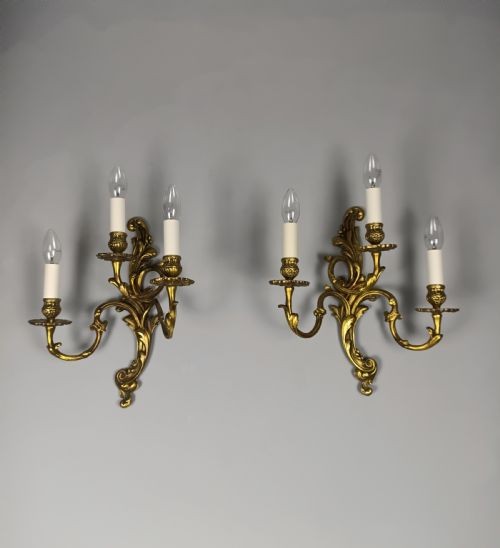 A pair of ornate gilded brass wall lights featuring three curving candle arms and Rococo scrollwork.