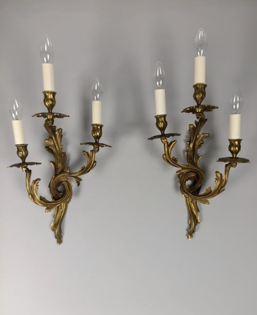 A pair of French Rococo style gilded bronze triple-arm wall sconces featuring ornate acanthus leaf scrollwork and candle-effect bulbs.