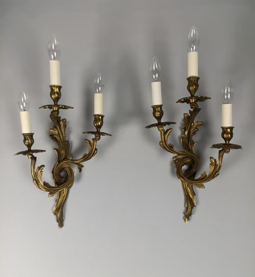 A pair of French Rococo style gilded bronze triple-arm wall sconces featuring ornate acanthus leaf scrollwork and candle-effect bulbs. View III