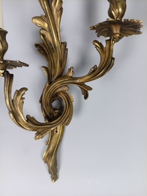 A pair of French Rococo style gilded bronze triple-arm wall sconces featuring ornate acanthus leaf scrollwork and candle-effect bulbs. View V