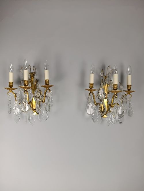 A pair of French Baroque style wall sconces featuring gilded brass frames with three arms each and hanging cut crystal drops.
