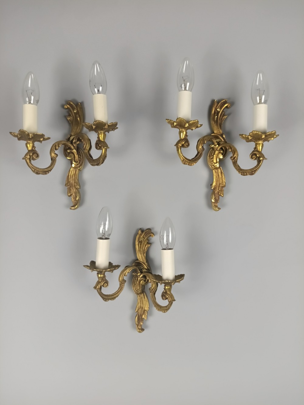 A set of three French Rococo style gilded metal twin-arm wall lights with scrollwork detailing and candle bulb fittings.