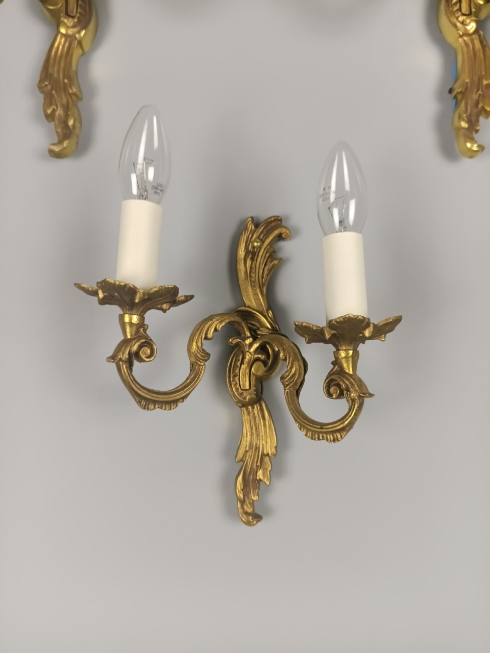 A set of three French Rococo style gilded metal twin-arm wall lights with scrollwork detailing and candle bulb fittings. View V