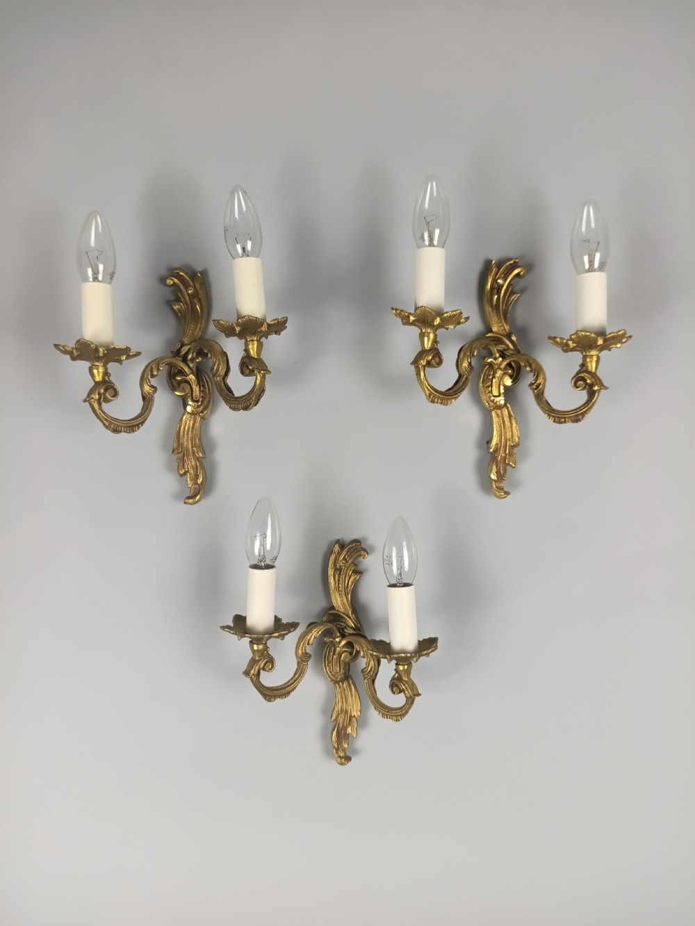 A set of three French Rococo style gilded metal twin-arm wall lights with scrollwork detailing and candle bulb fittings. View VI