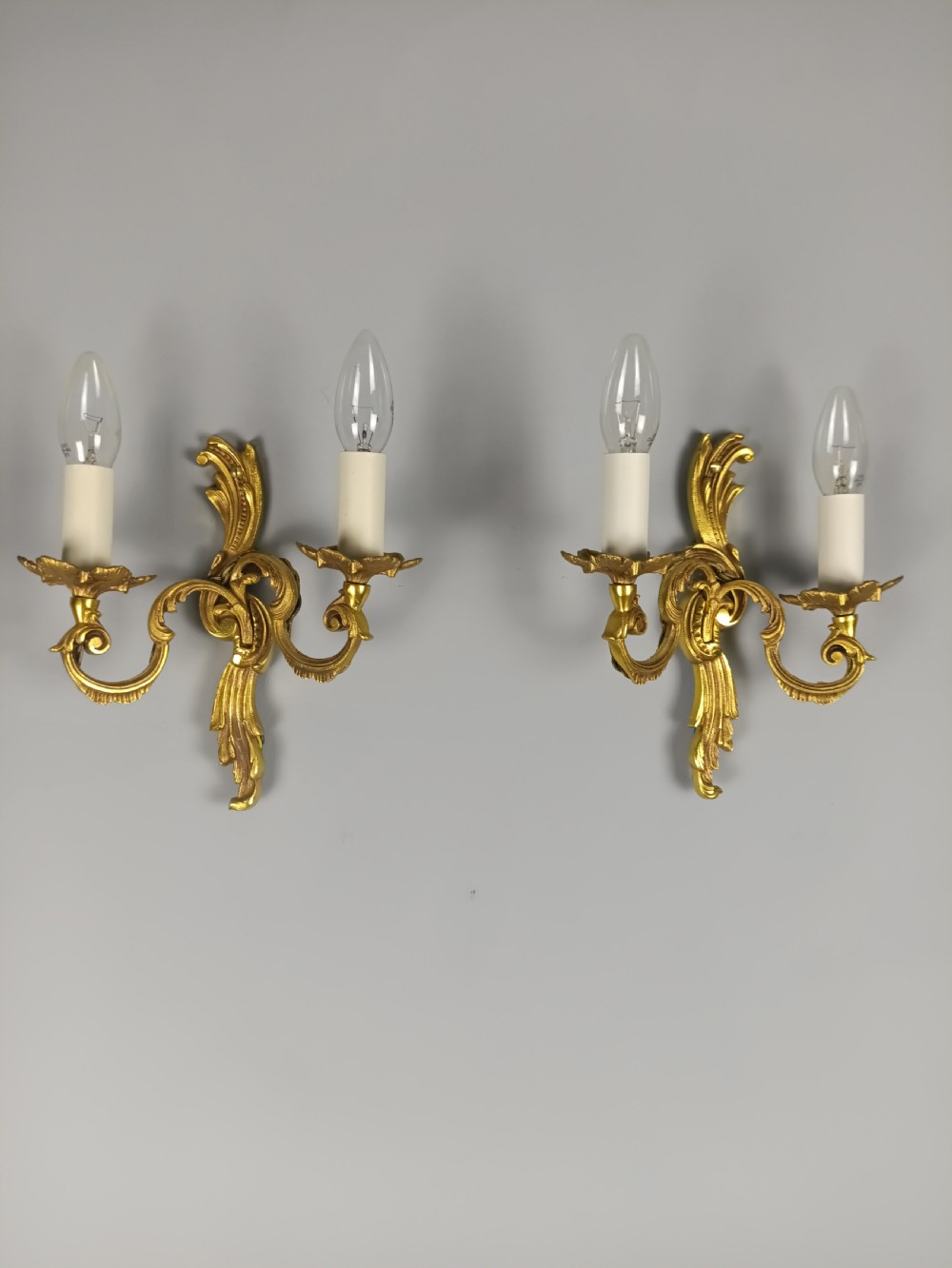 A pair of French Rococo style gilded metal twin-arm wall sconces featuring acanthus leaf scrollwork and candle-style holders.