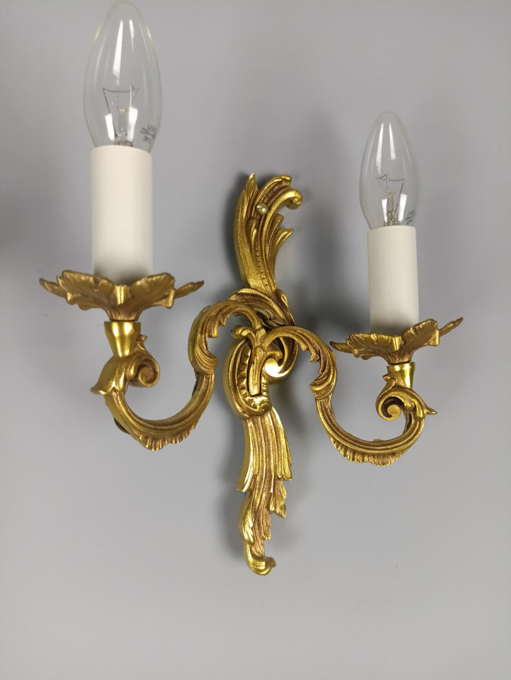 A pair of French Rococo style gilded metal twin-arm wall sconces featuring acanthus leaf scrollwork and candle-style holders. View V