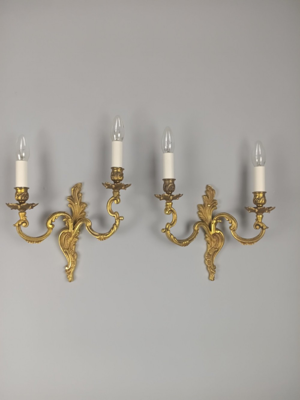 A pair of antique French Rococo gilded bronze twin-arm wall sconces featuring ornate acanthus leaf scrolling and candle sockets.
