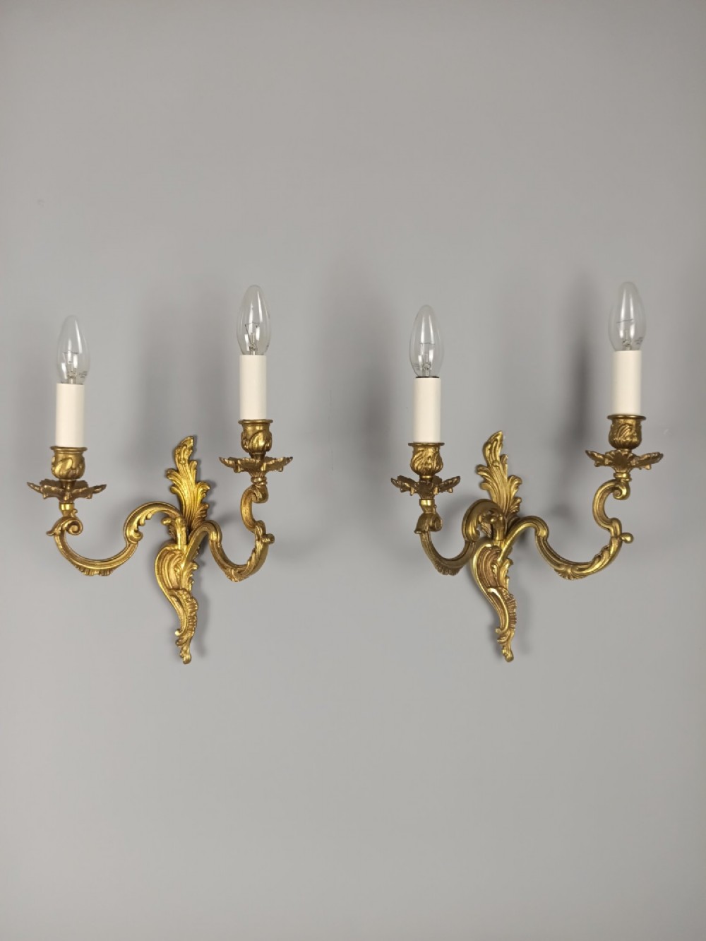 A pair of antique French Rococo gilded bronze twin-arm wall sconces featuring acanthus leaf motifs and candle sleeve light fixtures.