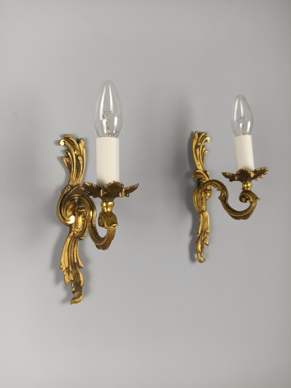 A pair of French Rococo style gilded bronze single-arm wall sconces featuring acanthus leaf scrollwork and candle-form sockets.