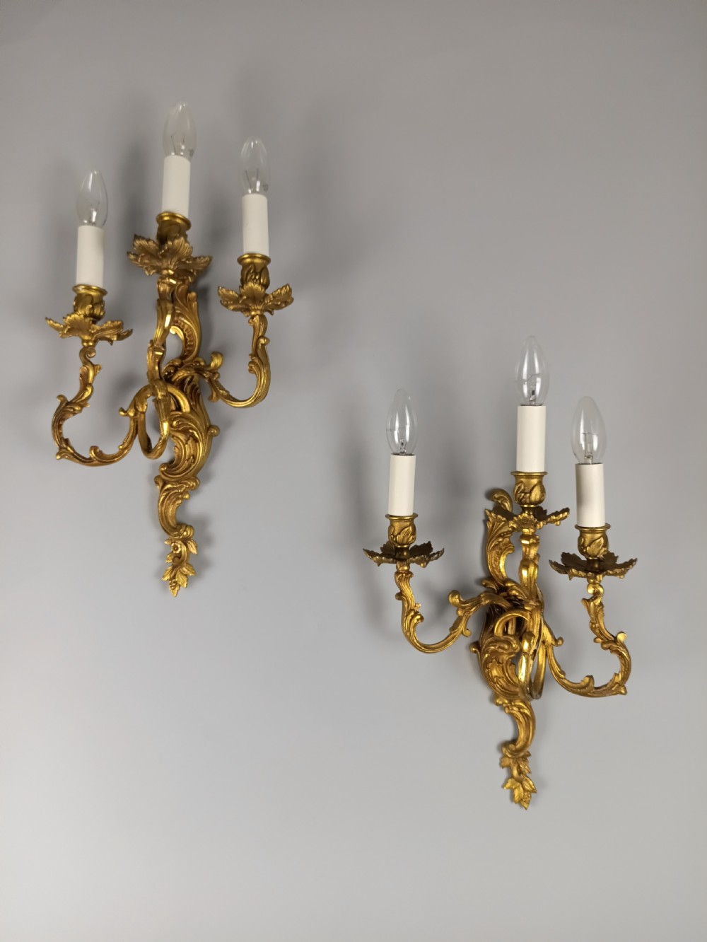 A pair of French Rococo style gilded bronze wall lights featuring three scrolling foliate arms and leaf motifs.