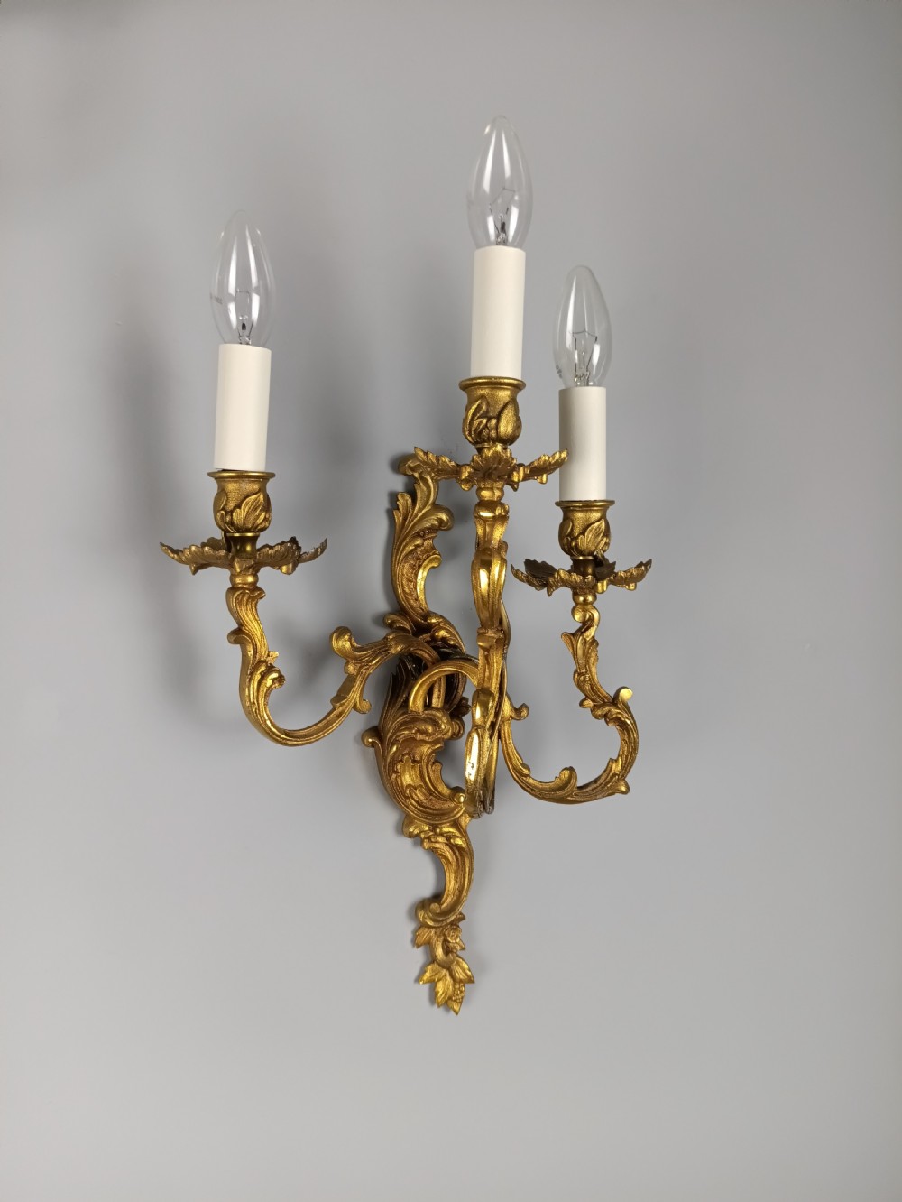 A pair of French Rococo style gilded bronze wall lights featuring three scrolling foliate arms and leaf motifs. View IV