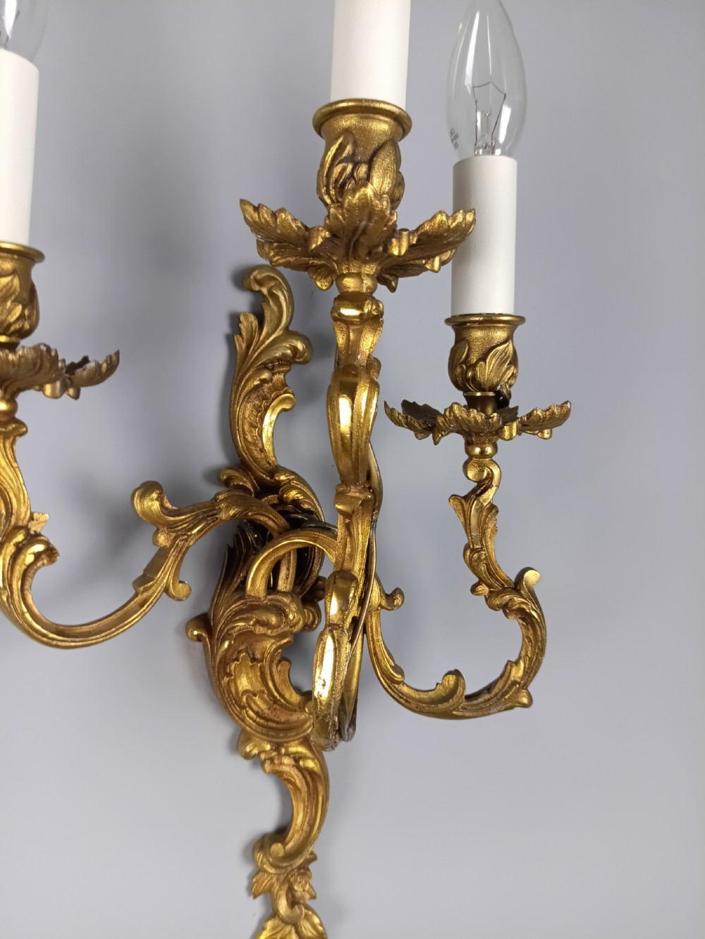A pair of French Rococo style gilded bronze wall lights featuring three scrolling foliate arms and leaf motifs. View V