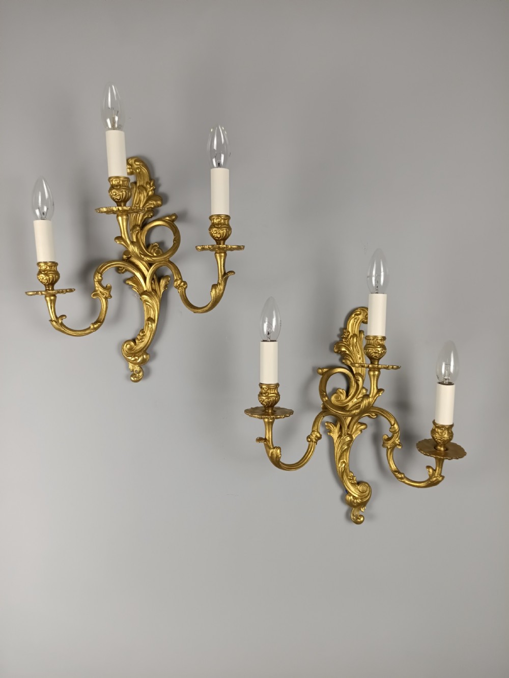 A pair of French Rococo style gilded brass triple arm wall sconces featuring ornate acanthus leaf scrollwork and candle shaped light fittings.