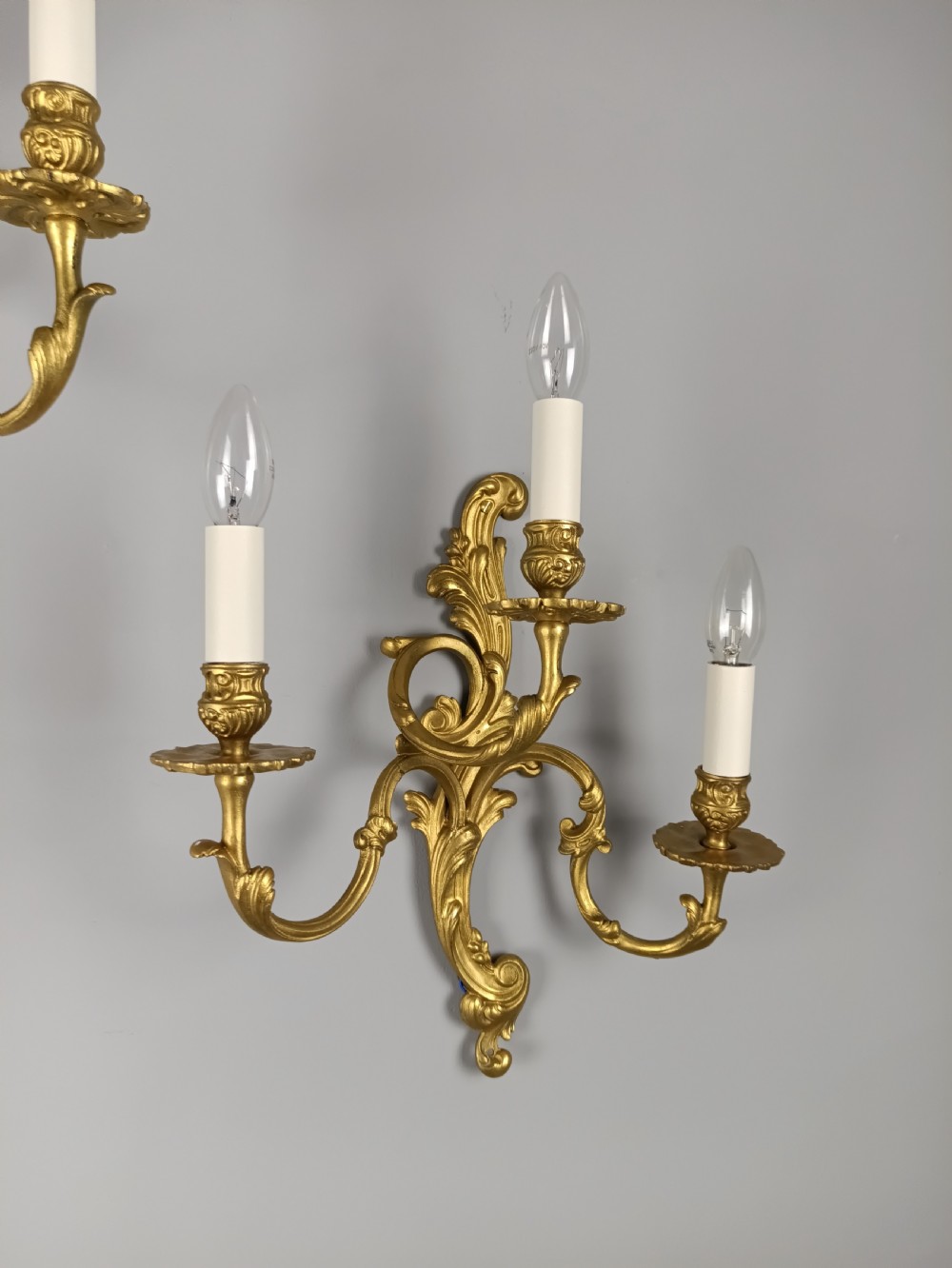 A pair of French Rococo style gilded brass triple arm wall sconces featuring ornate acanthus leaf scrollwork and candle shaped light fittings. View II