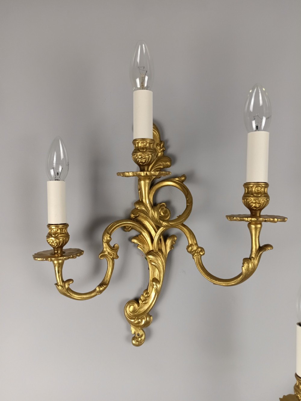 A pair of French Rococo style gilded brass triple arm wall sconces featuring ornate acanthus leaf scrollwork and candle shaped light fittings. View III
