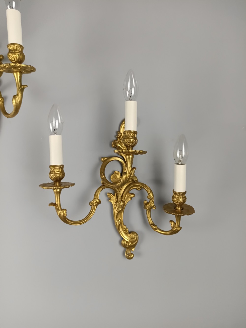 A pair of French Rococo style gilded brass triple arm wall sconces featuring ornate acanthus leaf scrollwork and candle shaped light fittings. View V