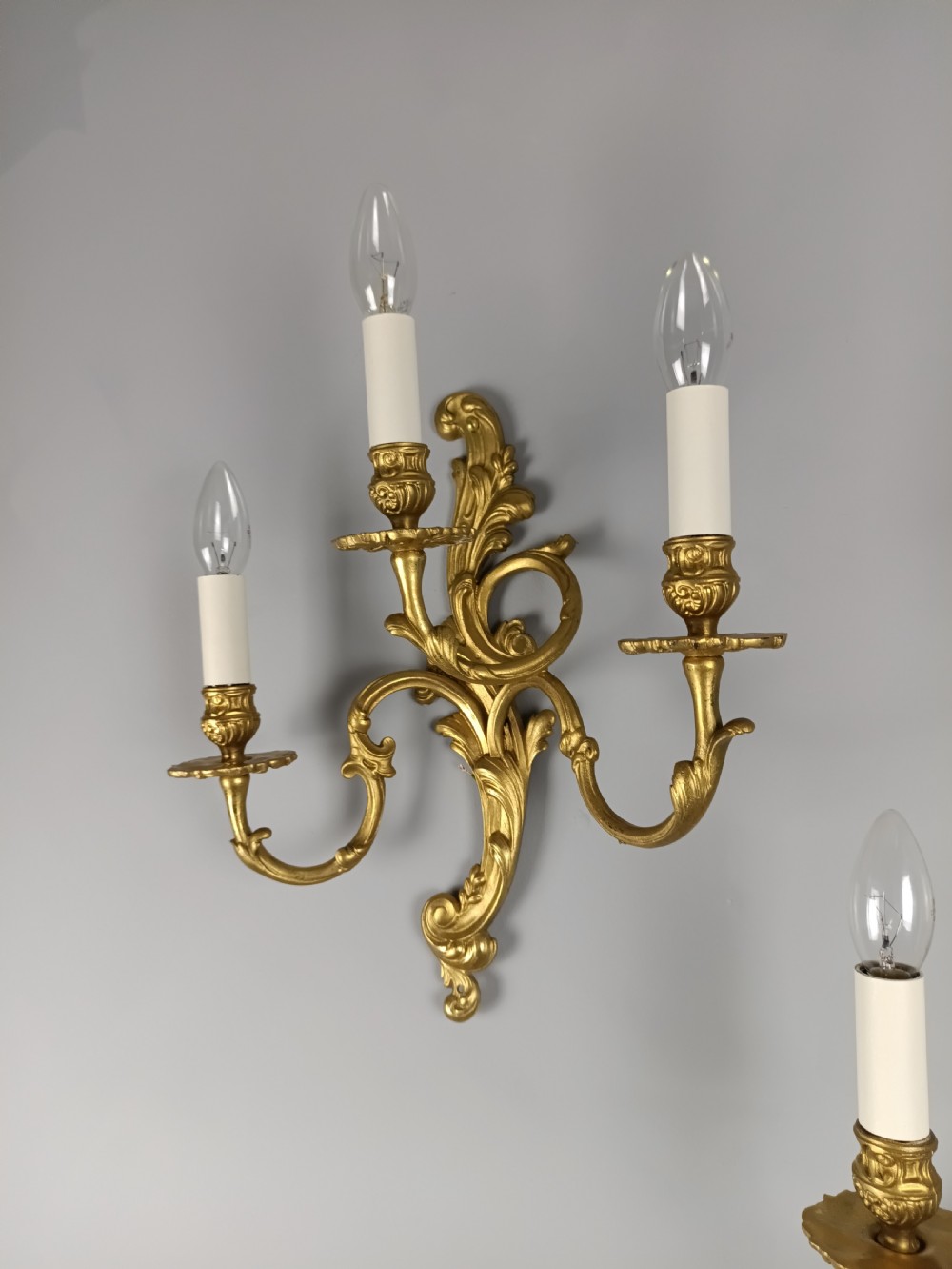 A pair of French Rococo style gilded brass triple arm wall sconces featuring ornate acanthus leaf scrollwork and candle shaped light fittings. View VI