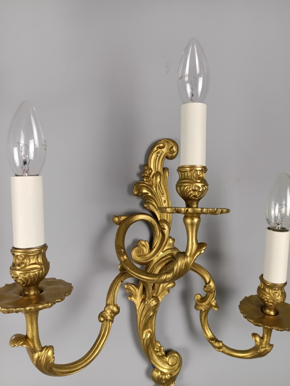 A pair of French Rococo style gilded brass triple arm wall sconces featuring ornate acanthus leaf scrollwork and candle shaped light fittings. View VIII