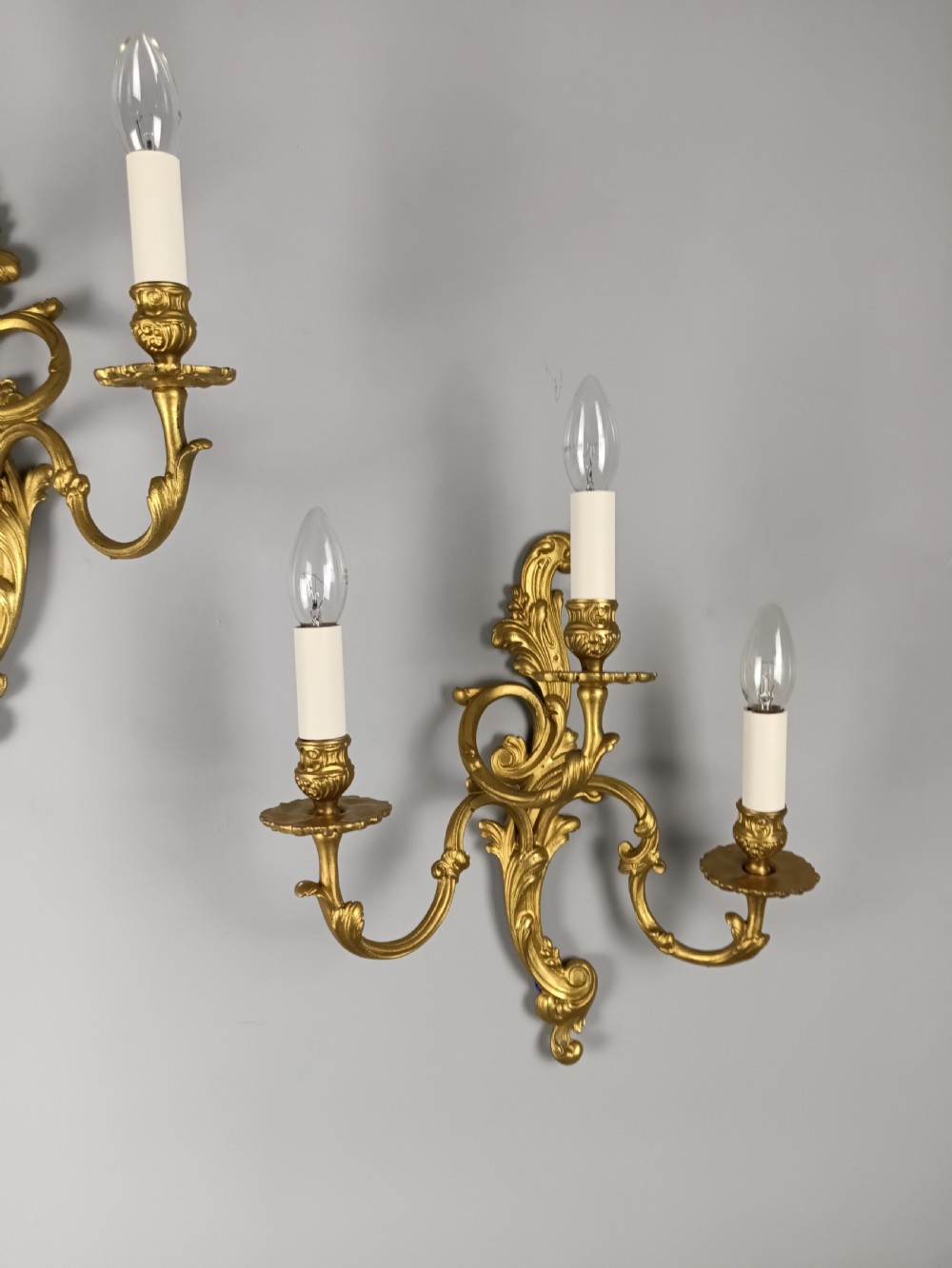 A pair of French Rococo style gilded brass triple arm wall sconces featuring ornate acanthus leaf scrollwork and candle shaped light fittings. View IX
