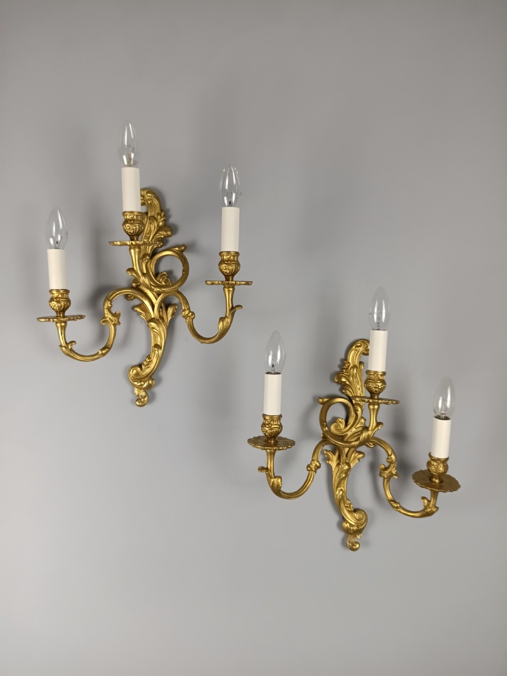 A pair of French Rococo style gilded brass triple arm wall sconces featuring ornate acanthus leaf scrollwork and candle shaped light fittings. View X