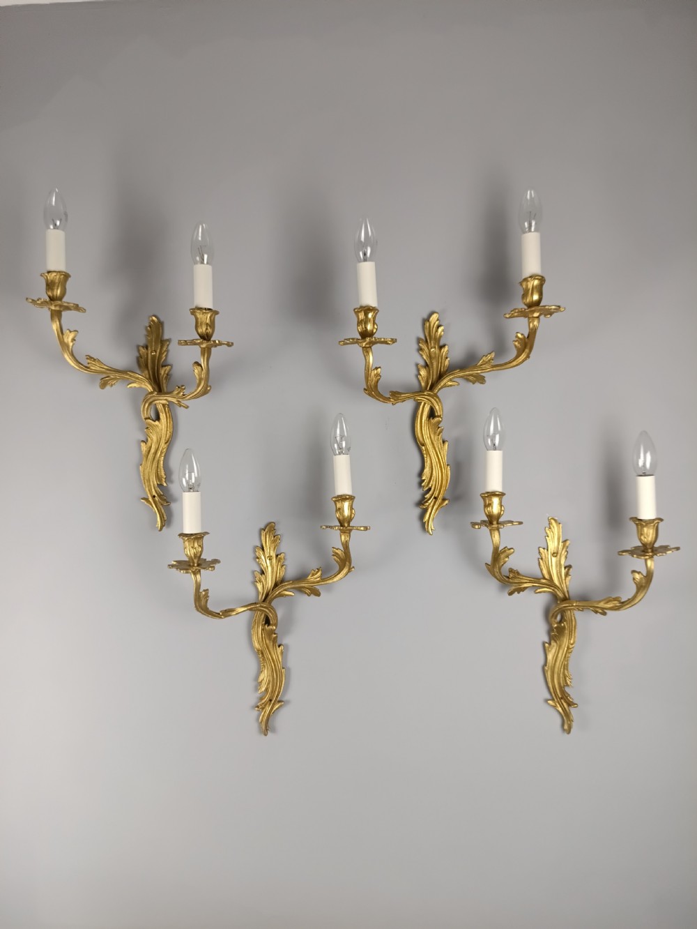 A set of four antique French Rococo style gilt bronze wall lights featuring twin curving foliate arms and acanthus leaf backplates.