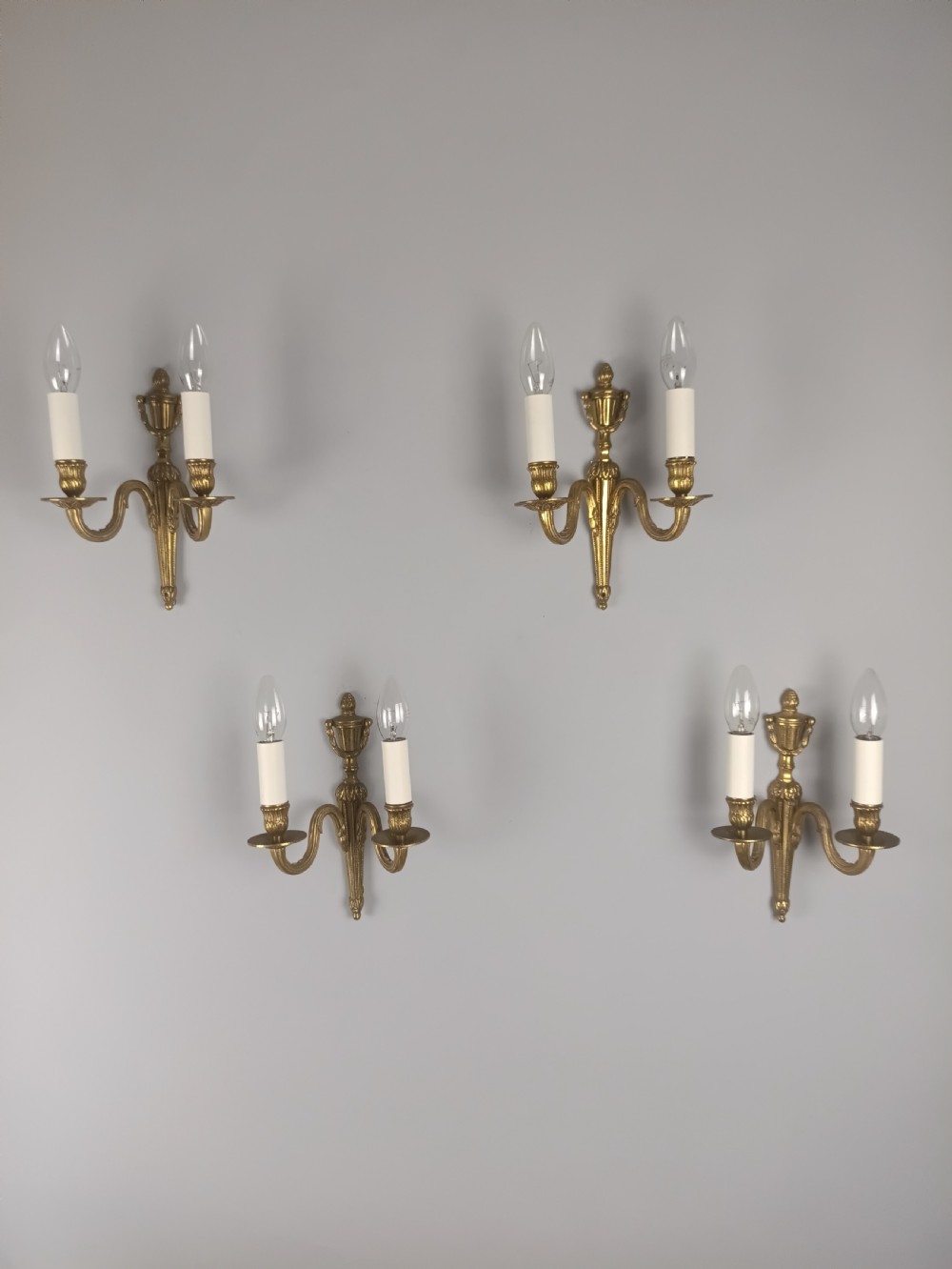A set of four French antique twin-arm wall lights crafted from gilded brass with neoclassical urn backplates and faux candle sleeves.