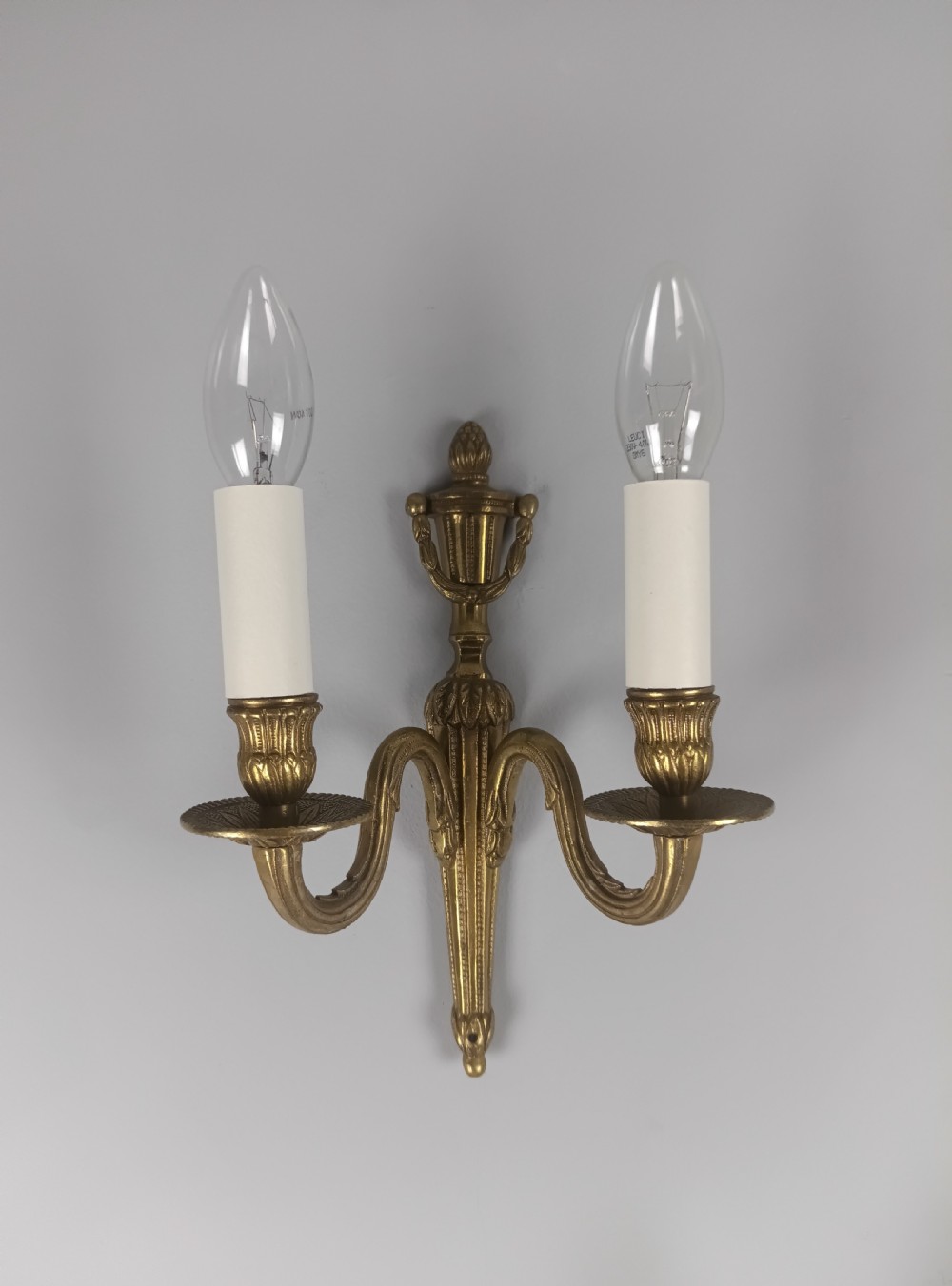 A set of four French antique twin-arm wall lights crafted from gilded brass with neoclassical urn backplates and faux candle sleeves. View V