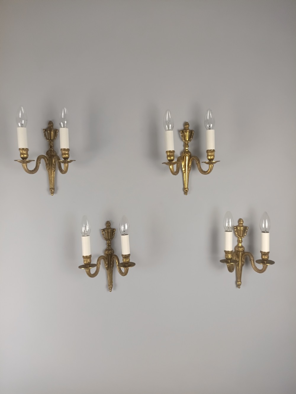 A set of four French antique twin-arm wall lights crafted from gilded brass with neoclassical urn backplates and faux candle sleeves. View X
