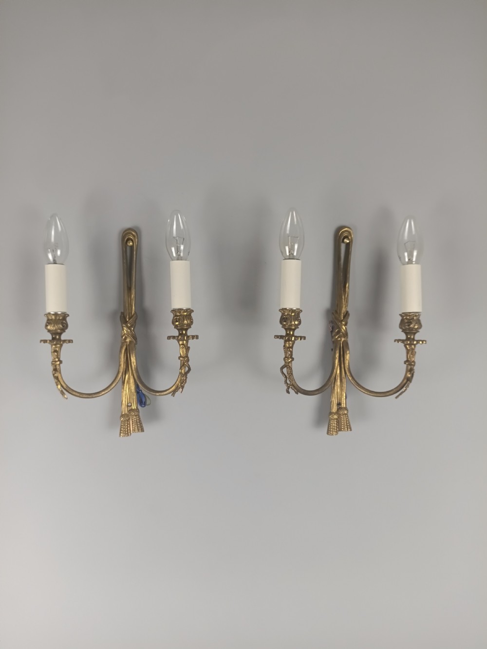 A pair of antique French gilt brass wall sconces featuring ribbon bow top motifs, tassel backplates, and twin candle arms.