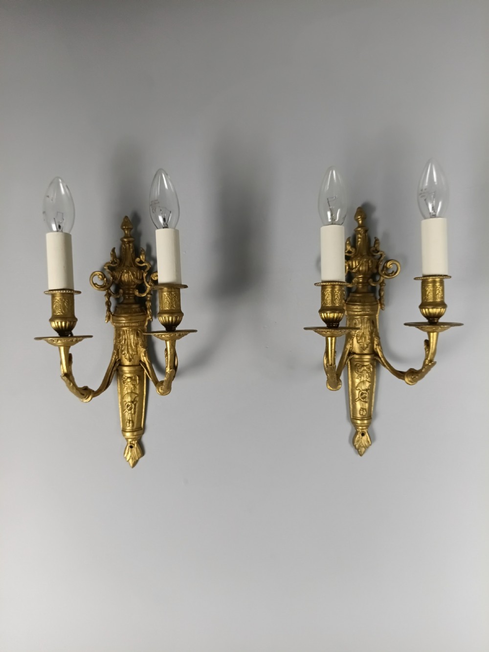 A pair of 19th-century French gilt brass twin-arm wall sconces featuring urn-topped backplates and classical foliage relief work.