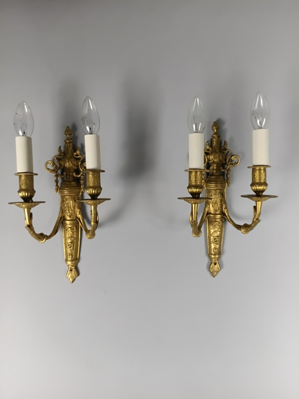 A pair of 19th-century French gilt brass twin-arm wall sconces featuring urn-topped backplates and classical foliage relief work. View V