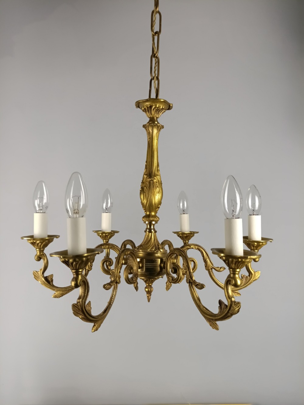 A six-arm French gilded brass chandelier featuring ornate acanthus leaf embellishments, candle-style bulb holders, and an articulated central column.