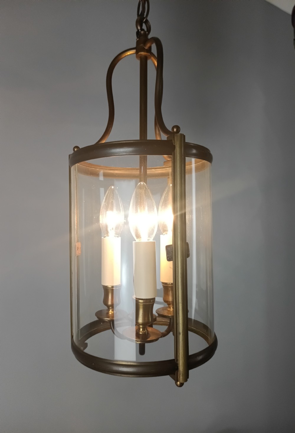 A cylindrical gilded brass hall lantern featuring curved clear glass panels and an internal three-light candle cluster. View X