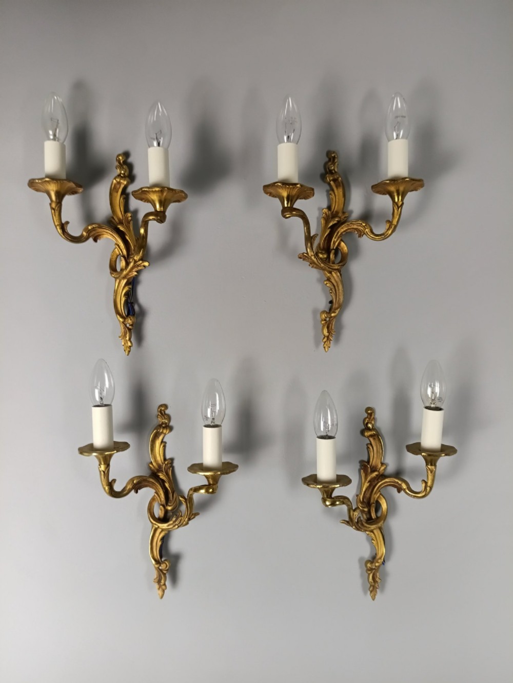 Four French Rococo style gilded bronze twin-arm wall lights with foliate scrollwork and white candle sleeves.