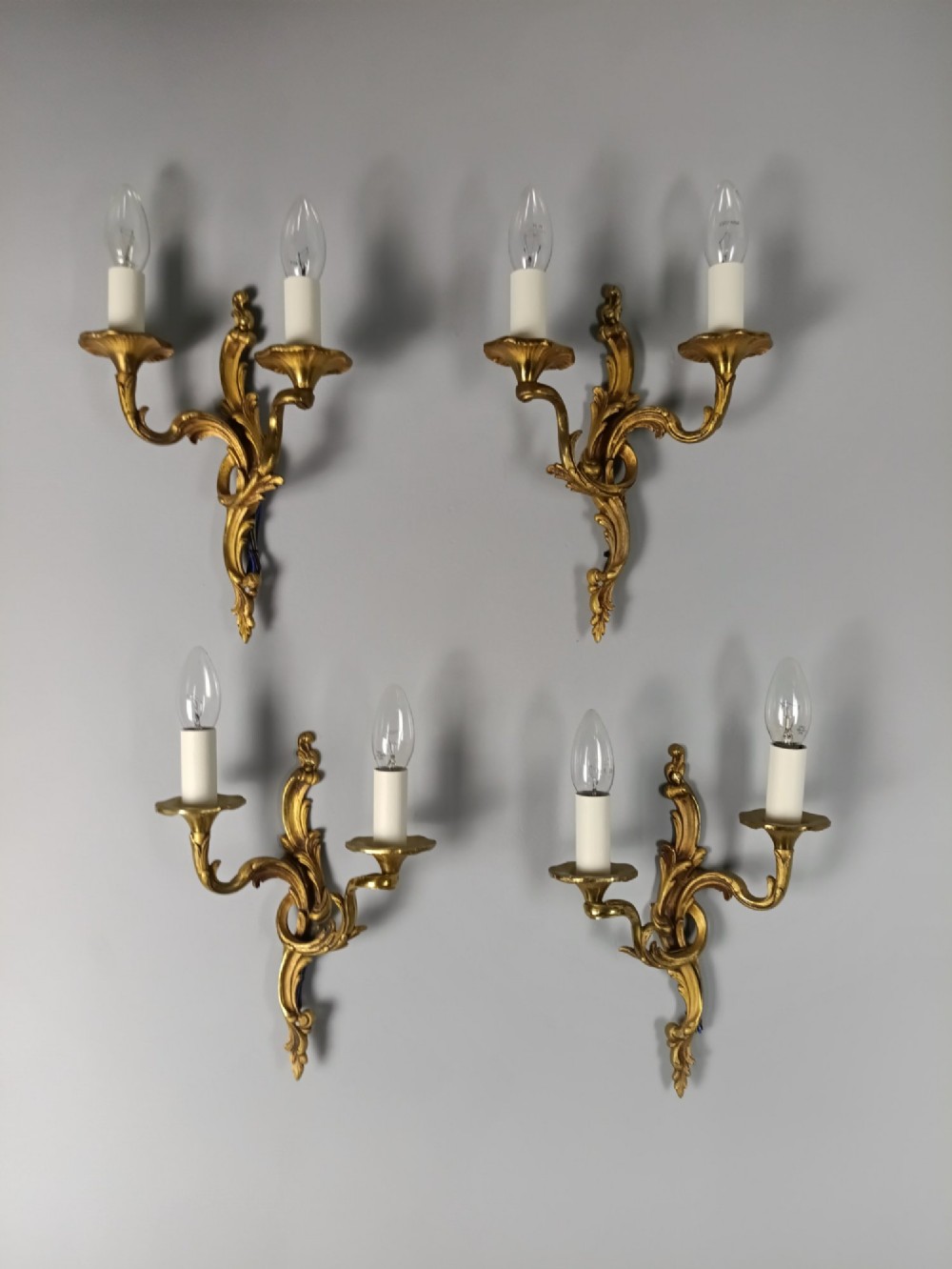 Four French Rococo style gilded bronze twin-arm wall lights with foliate scrollwork and white candle sleeves. View VIII