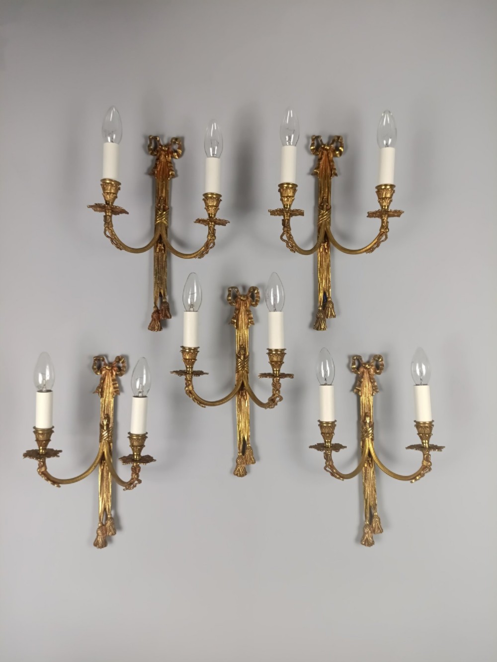 A set of five French gilded brass twin-arm wall sconces featuring ribbon bow crests, draped tassel detailing, and candle-style bulb holders.