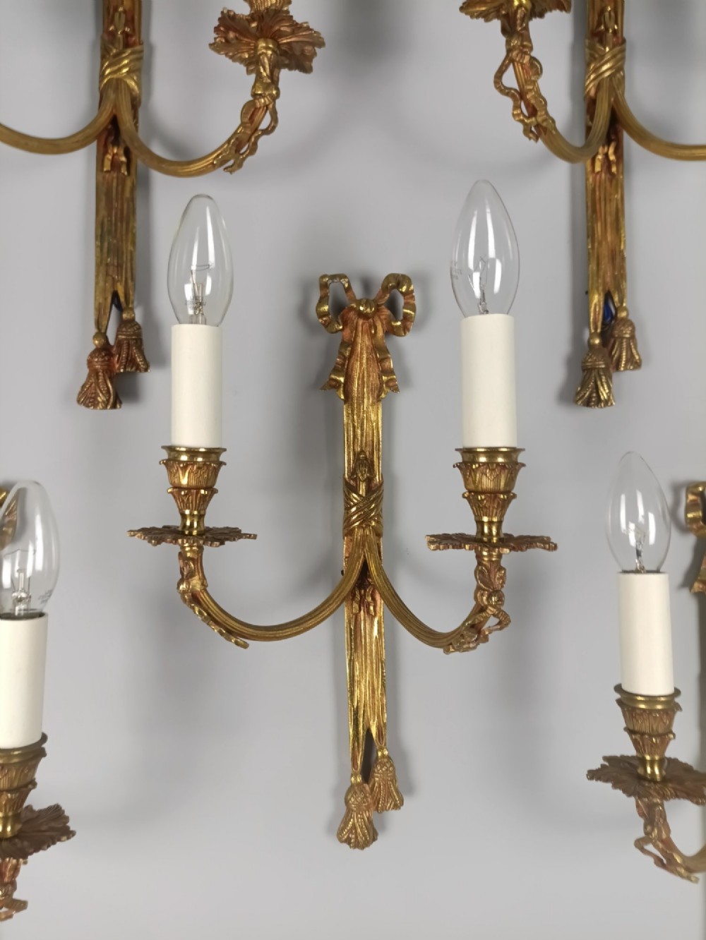 A set of five French gilded brass twin-arm wall sconces featuring ribbon bow crests, draped tassel detailing, and candle-style bulb holders. View IV