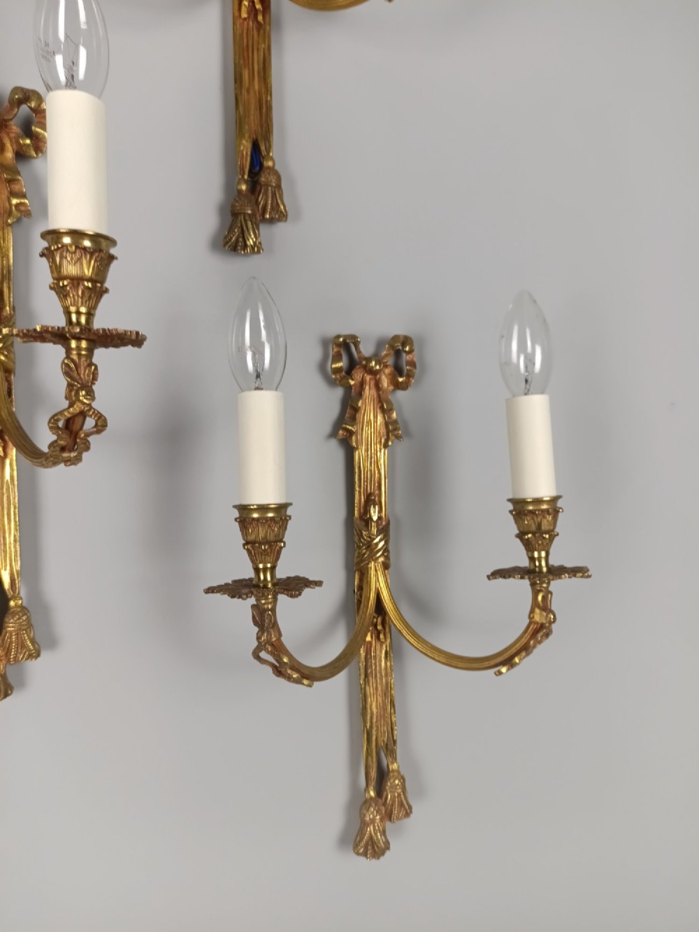 A set of five French gilded brass twin-arm wall sconces featuring ribbon bow crests, draped tassel detailing, and candle-style bulb holders. View VI