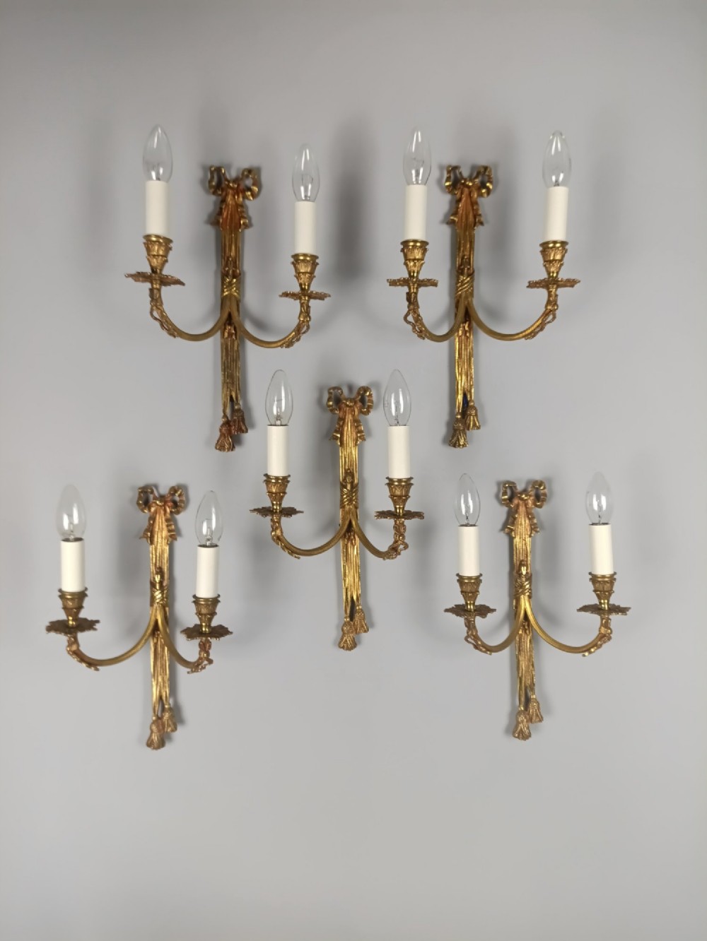 A set of five French gilded brass twin-arm wall sconces featuring ribbon bow crests, draped tassel detailing, and candle-style bulb holders. View VII