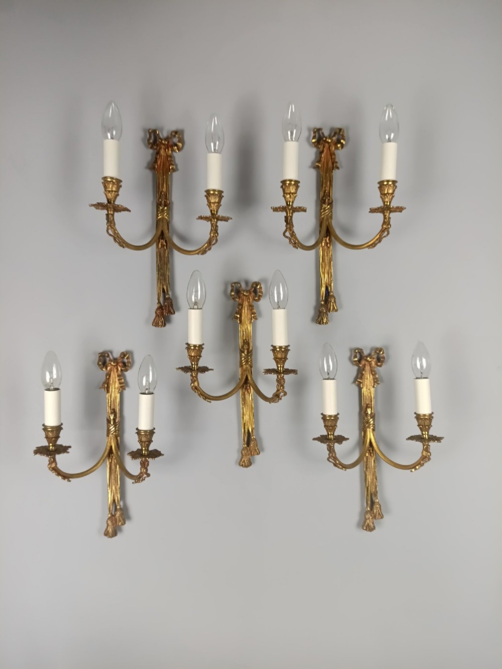 A set of five French gilded brass twin-arm wall sconces featuring ribbon bow crests, draped tassel detailing, and candle-style bulb holders. View VIII