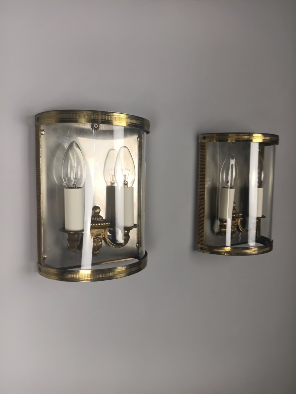 A pair of French gilt brass half-lantern wall lights featuring curved clear glass panels and twin candle-style sockets.