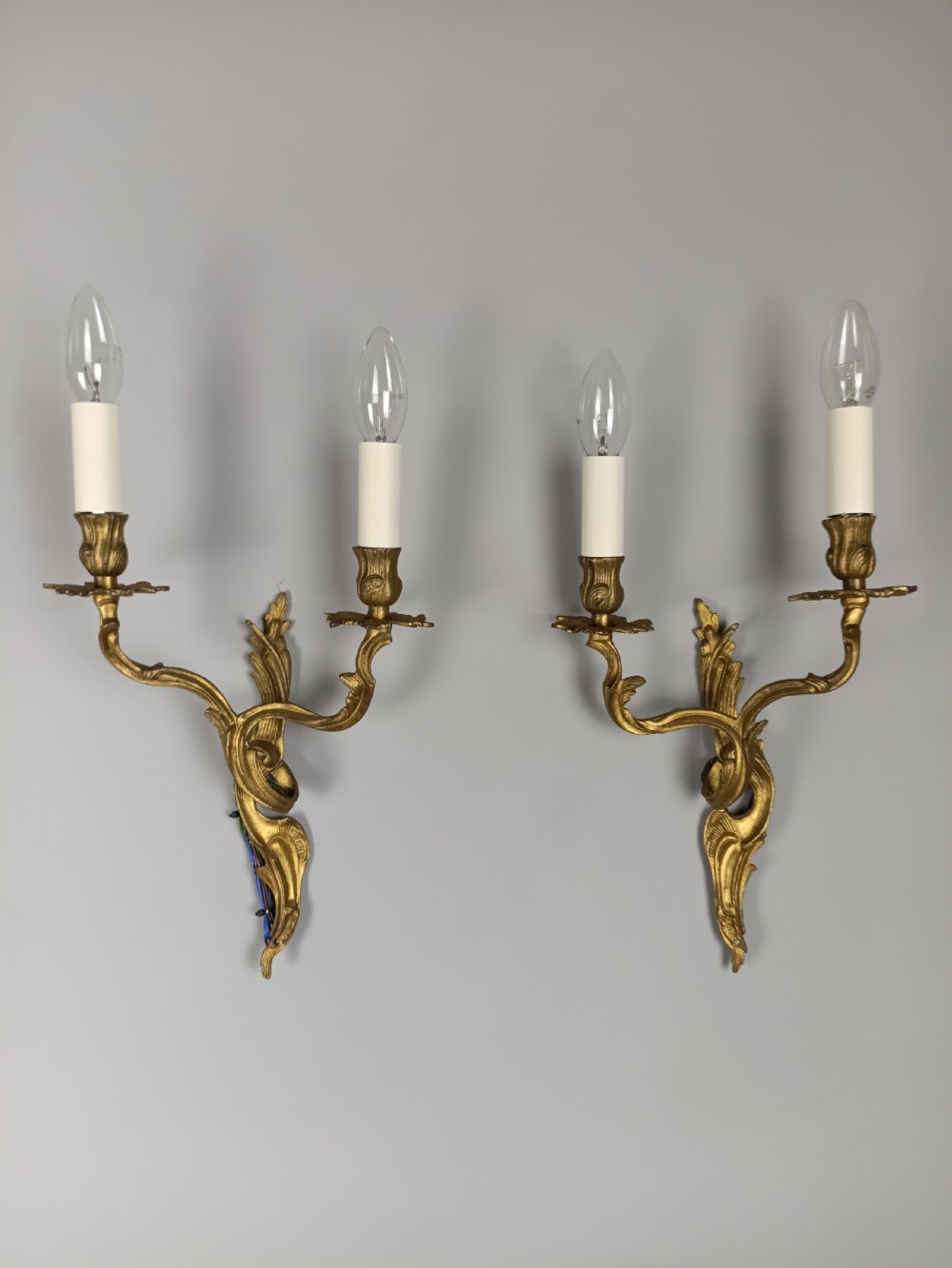 A pair of two-arm gilded brass wall sconces featuring ornate Rococo scrolling leaf details and white candle-effect sleeves.