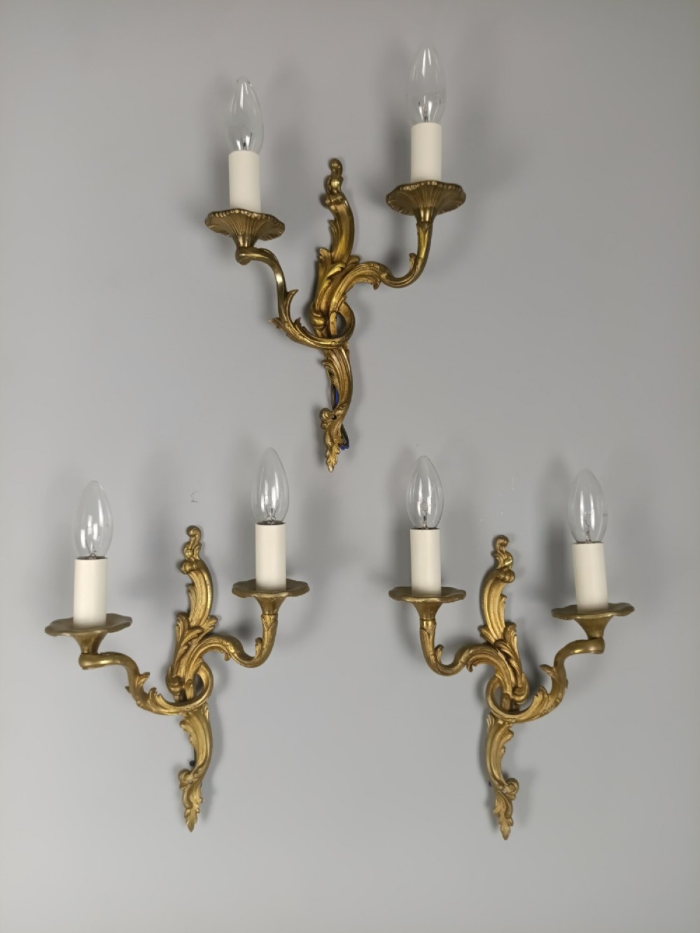 A set of three French gilded bronze twin-arm wall lights with ornate foliage scrolling and candle-style sockets.