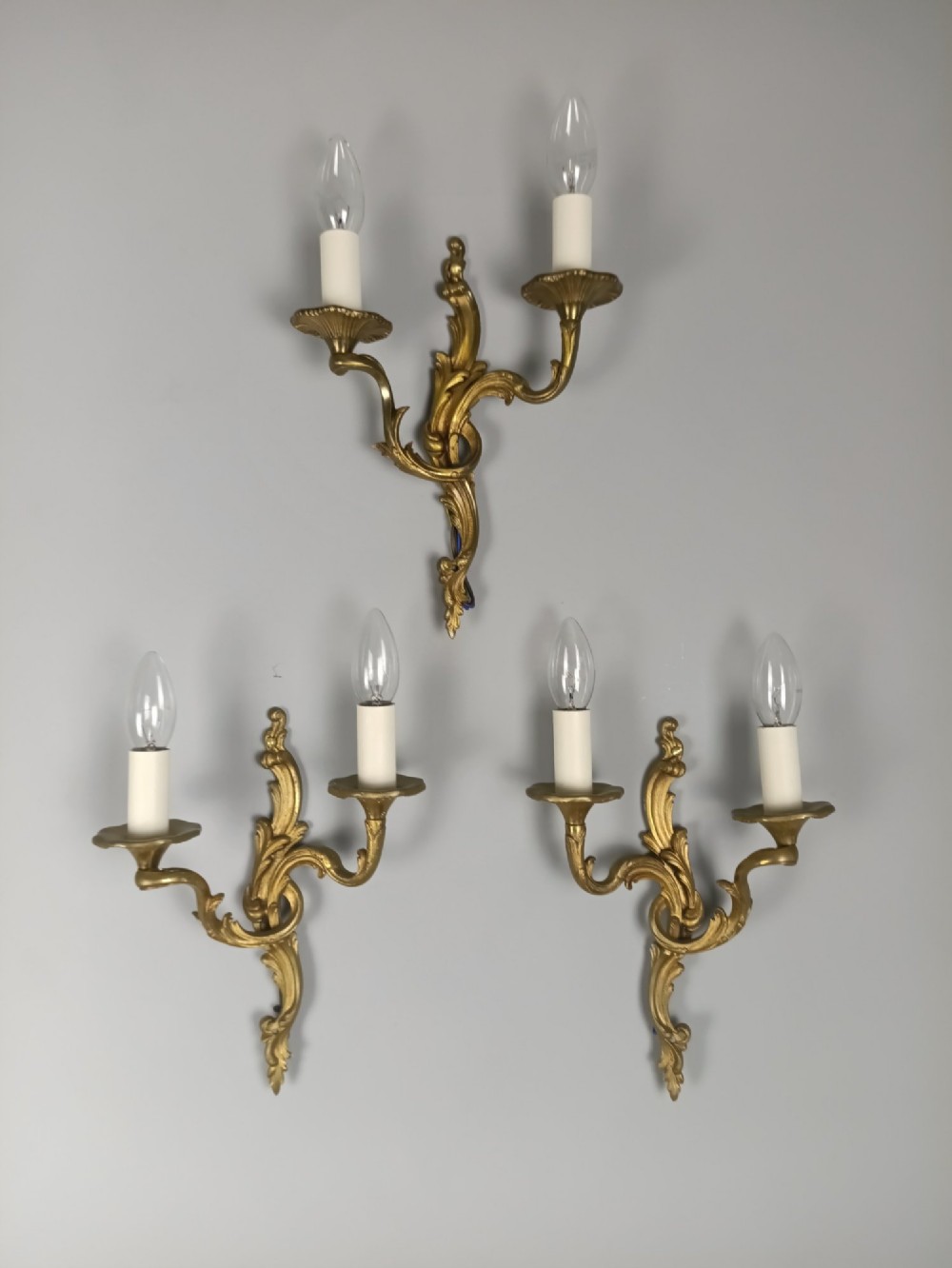 A set of three French gilded bronze twin-arm wall lights with ornate foliage scrolling and candle-style sockets. View VI