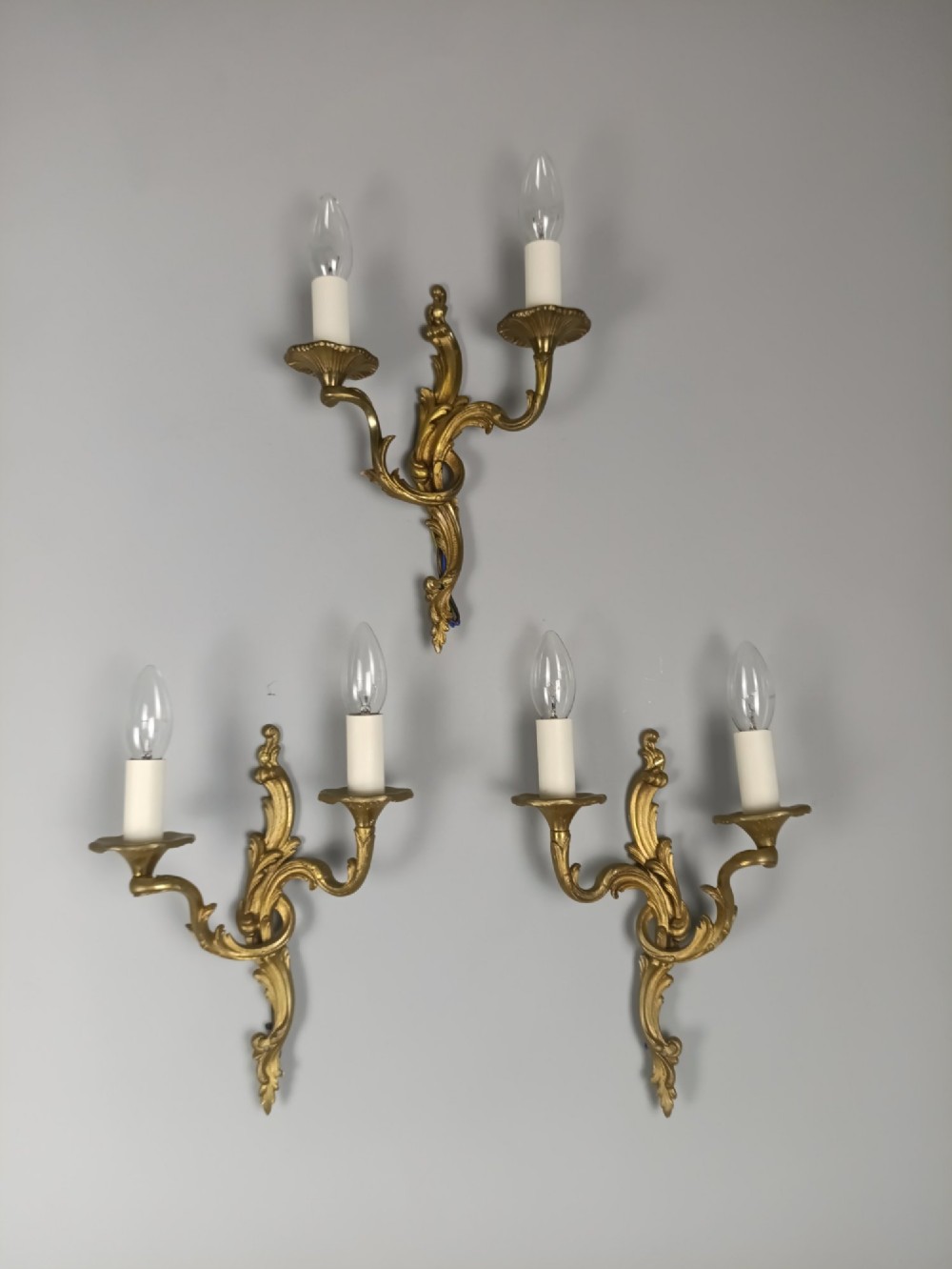A set of three French gilded bronze twin-arm wall lights with ornate foliage scrolling and candle-style sockets. View IX